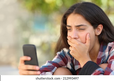 Worried Teenager Girl Looking At Her Smart Phone In A Park With An Unfocused Background