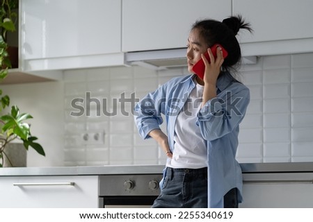 Worried upset Asian woman housewife standing alone in kitchen making unpleasant phone call, talking on smartphone with concerned face expression. Sad mother calling child who ignoring parent calls
