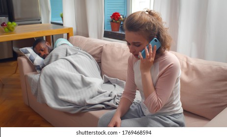 Worried Wife Calling Doctor From Home For Husband Got Sick Lying On Couch Covered In Towel And Coughing. Young Woman Consulting With Doctor On Phone About Ill Boyfriend