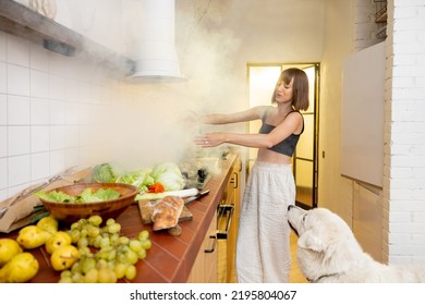 Worried Woman Has A Problem Preparing Food, Having Burnt Meal On Kitchen Hob. Confused Housewife Waves Her Hands With Her Dog In The Smoke