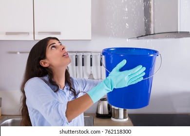 Worried Woman Holding Bucket While Water Droplets Leak From Ceiling In Kitchen
