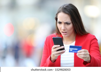 Worried woman holding credit card looking at smart phone in the street