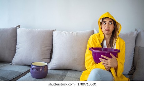 Worried Woman in Raincoat Holding Bucket While Water Droplets Leak From Ceiling in Living Room. Woman Holding Bucket While Water Droplets Leak From Ceiling. Roof Leaking 
