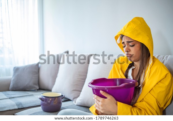 Worried Woman in\
Raincoat Holding Bucket While Water Droplets Leak From Ceiling in\
Living Room. Woman Holding Bucket While Water Droplets Leak From\
Ceiling. Roof Leaking 