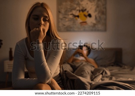 worried woman sitting on bed near blurred boyfriend using smartphone in bedroom
