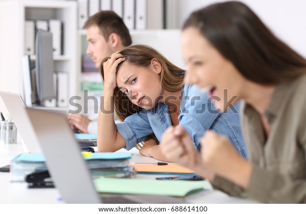Worried worker beside a successful one who\
is excited reading good news on line at\
office