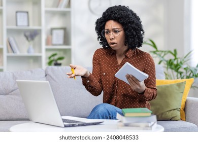 Worried young African American female student studying at home by distance learning, holding notebook in hand and looking frustrated at laptop screen. - Powered by Shutterstock