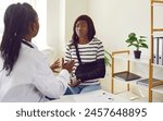 Worried young African American woman patient with black sling on broken arm sitting at table and talking to professional doctor traumatologist about treatment of her injury