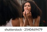 Worried young beautiful African American woman sad ethnic girl looking in mirror stressed about facial wrinkles problem acne frustrated upset touching face dry sensitive skin care facial blackheads