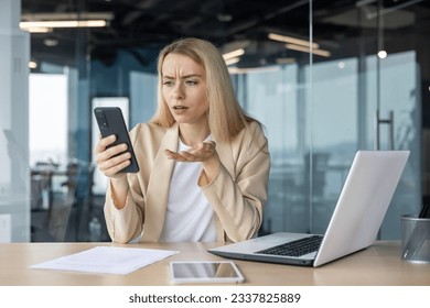 Worried young business woman working in the office at the laptop, holding the phone in her hands, anxiously looking at the screen.