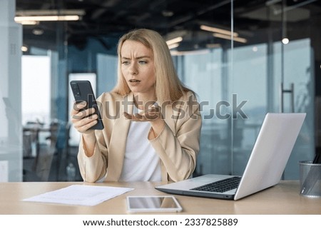 Worried young business woman working in the office at the laptop, holding the phone in her hands, anxiously looking at the screen.