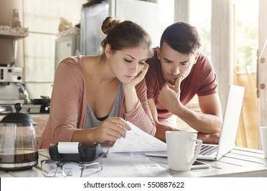 Worried young Caucasian married couple reading important notification from bank while managing domestic finances and calculating their expenses at kitchen table, using laptop computer and calculator