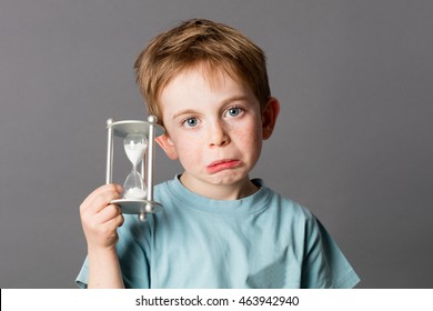 Worried Young Child With Sad Blue Eyes And A Pouting Mouth Holding An Egg Timer, Showing His Disappointment In Growing Up For Time Concept, Grey Background