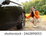 Worried young European woman talking on cellphone and checking her car flat tire on the side of the highway, calling for help