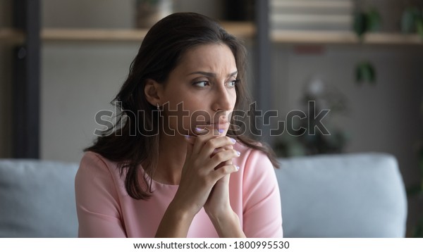 Worried young indian Arabic woman sit on couch at\
home look in distance thinking pondering, anxious unhappy arab\
mixed race female suffer from mental psychological personal\
problems, mourn or\
yearn
