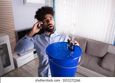 Worried Young Man Calling Plumber While Leakage Water Falling Into Bucket At Home