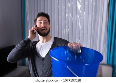 Worried Young Man Calling Plumber While Leakage Water Falling Into Bucket At Home