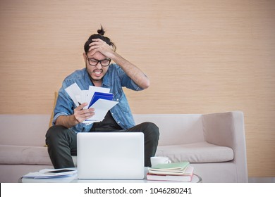 Worried Young Man Paying His Bills Online With Laptop In The Living Room At Home. Nerd Man At Desk With Computer And Checkbook, Worrying About Paying Bills, Holding His Head.