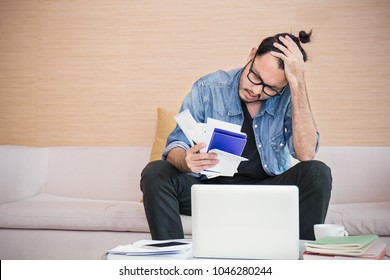 Worried Young Man Paying His Bills Online With Laptop In The Living Room At Home. Nerd Man At Desk With Computer And Checkbook, Worrying About Paying Bills, Holding His Head.