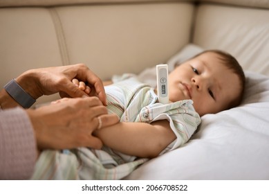 Worried Young Mother Sitting On Sofa Beside Her Sick Son With High Fever. Mom Measures Temperature Using Thermometer Of Sick Child Lying Under Blanket At Home.