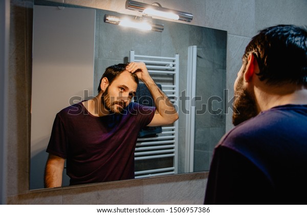 A worried young white man looks at himself in
the mirror and inspects his premature receding hairline. Attractive
Caucasian male adult in his 30s concerned about losing hair. Male
pattern baldness