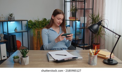 Worried Young Woman Businesswoman Enthusiastically Playing Racing Or Shooter Video Games On Smartphone Sitting At Table Working From Home Office. Girl Freelancer Study, E-learning, Remote Job Online