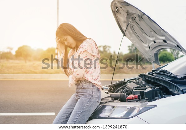 Worried Young woman holding his head she opened the\
hood Broken car on the side See engines that are damaged with\
raised hood on the road