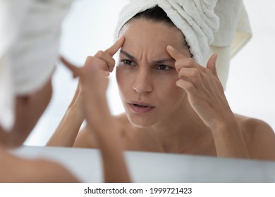 Worried young woman wrapped in towel looking in mirror in bathroom, finding skin defects, touching forehead, discovering pimples, acne, wrinkles. Facial skin problems, cosmetology concept