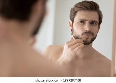 Worry Young Handsome Man Examining Face In Mirror Reflection, Touching Chin And Stubble With Concerned Face, Discovering Dryness, Wrinkles, Skin Problems, Feeling Discomfort. Male Beauty Care Concept