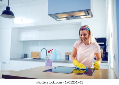 Worthy cheerful caucasian blond housewife with rubber gloves on hands cleaning stove. Kitchen interior.