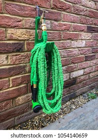 Wound Up Coiled Retractable Gree Garden Hose Hanging On A Plastic Hook Around A Brass Coloured Outdoor Tap On A Brick Wall.