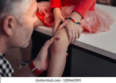 Wounded knee. Grey-haired tall father in a checkered shirt looking attentive while looking at his daughters wounded knee - Powered by Shutterstock