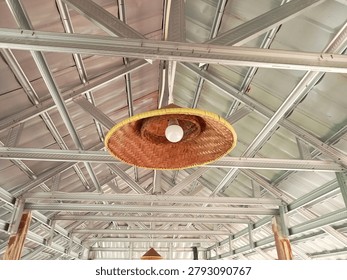 Woven bamboo hat repurposed as rustic pendant lamp hanging from metal roof truss in traditional Indonesian open-air pavilion.