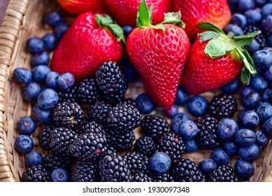 woven basket of red strawberries with green leafy tops, surrounded by ripe blackberries and blueberries