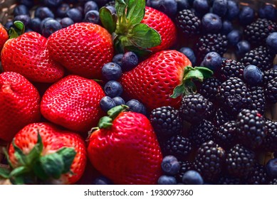 woven basket of red strawberries with green leafy tops, surrounded by ripe blackberries and blueberries