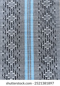 A woven fabric pattern with alternating black and white designs, featuring a repeated diamond-like motif. The design is framed by blue vertical stripes in the center, which adds contrast to the graysc