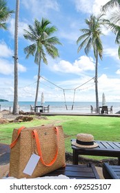 Woven Handbag And Hat Accessory For Holiday Put On Beach Bed With Background Green Field, Coconut Trees And Swing Hanging On, Blue Sea ,  White Clouds Blue Sky.