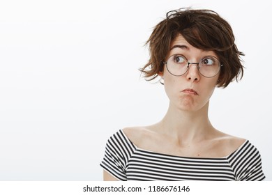 Wow, It Is Amazing But Unexpected. Impressed Cute And Funny Girl In Round Glasses With Stylish Short Haircut, Pulling Face In Not Bad Emotion, Looking Left Curiously, Standing Pleased Over Grey Wall