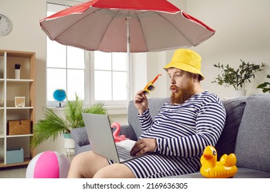 Wow, it's a bargain. Funny young man going on holiday, booking ticket online, sitting in hat and swimsuit on sofa with beach umbrella and inflatable toys, browsing best prices on travel agency website