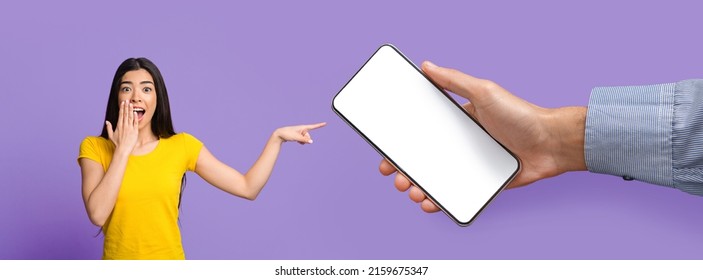 Wow, Check This Great Offer. Excited surprised Asian lady cover mouth point at big blank cell phone screen, huge hand holding device with white empty display on violet purple wall, banner panorama