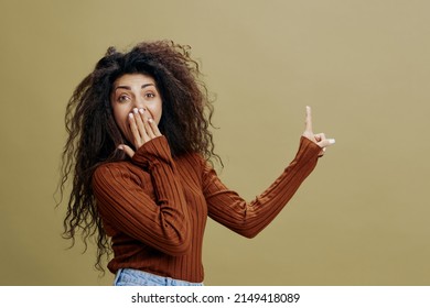 WOW CONCEPT. Shocked Pretty Curly Latin Woman Close Mouth, Look At Camera And Point Finger Up. Autumn Collection, Seasonal Sale Clothing Fashion Brands. Studio Shoot, Copy Space Green Background. 