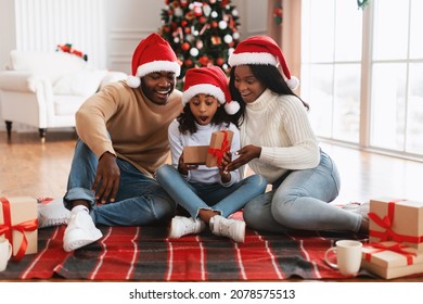 Wow. Cute excited black girl opening Christmas present box in festive living room. Smiling parents giving Xmas gift to surprised daughter sitting on floor blanket near decorated tree on holiday eve - Powered by Shutterstock