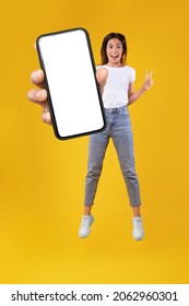 Wow. Emotional overjoyed young woman with excited funny facial expression holding big empty cell phone screen jumping up high in the air on orange studio background, showing peace sign gesture