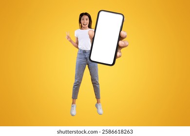 Wow. Emotional overjoyed young woman with excited funny facial expression holding big empty cell phone screen jumping up high in the air on orange studio background, showing peace sign gesture