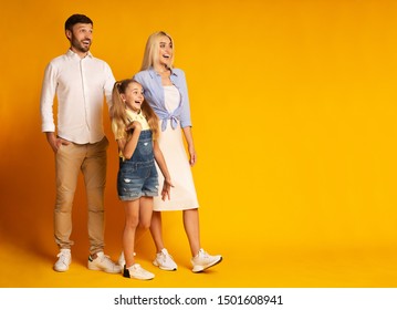 Wow. Excited Family Of Three Looking Aside At Empty Space Standing Together Over Yellow Background In Studio. Full Length