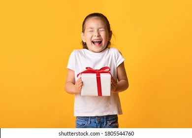 Wow. Excited Little Kid Holding Gift Box And Screaming With Joy, Closed Eyes And Open Mouth, Yellow Wall, Copyspace