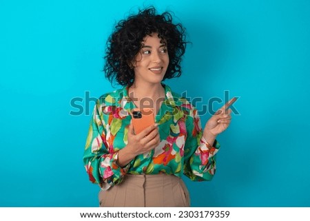 Wow!! excited young arab woman wearing colorful shirt over blue background showing mobile phone with open hand gesture