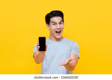 Wow Excited Young Asian Man Showing Mobile Phone With Open Hand Gesture Isolated On Yellow Background