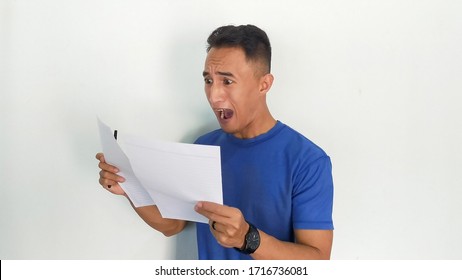 Wow Face Of Young Asian Man Shocked What He Look On The Paper While Read A Bill, Job, Or Letter. Indonesia Man Wear Blue Shirt Isolated White Background.