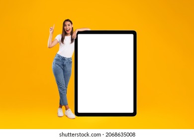 Wow, Great Offer, Cool Mobile App. Full Body Length Of Excited Lady Pointing Finger Up Leaning On Big Huge White Empty Tablet Pad Screen Standing On Bright Orange Studio Wall. Cell Display Mock Up Ad